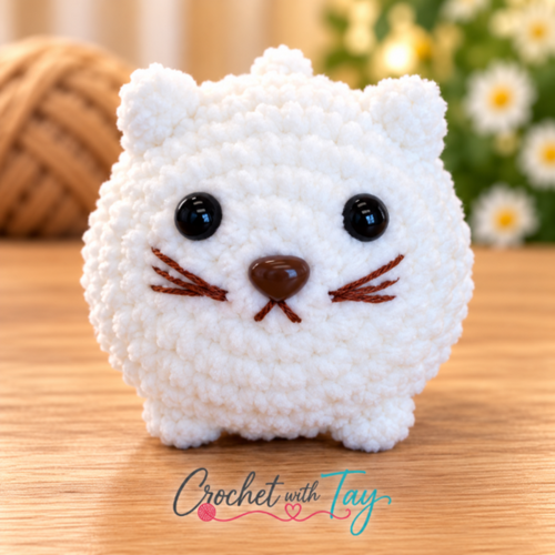A white crocheted cat-shaped plush toy with black bead eyes, a brown stitched nose, and brown whiskers, sitting on a wooden surface with a blurred yarn ball and flowers in the background. The text 'Crochet with Tay' is visible at the bottom.